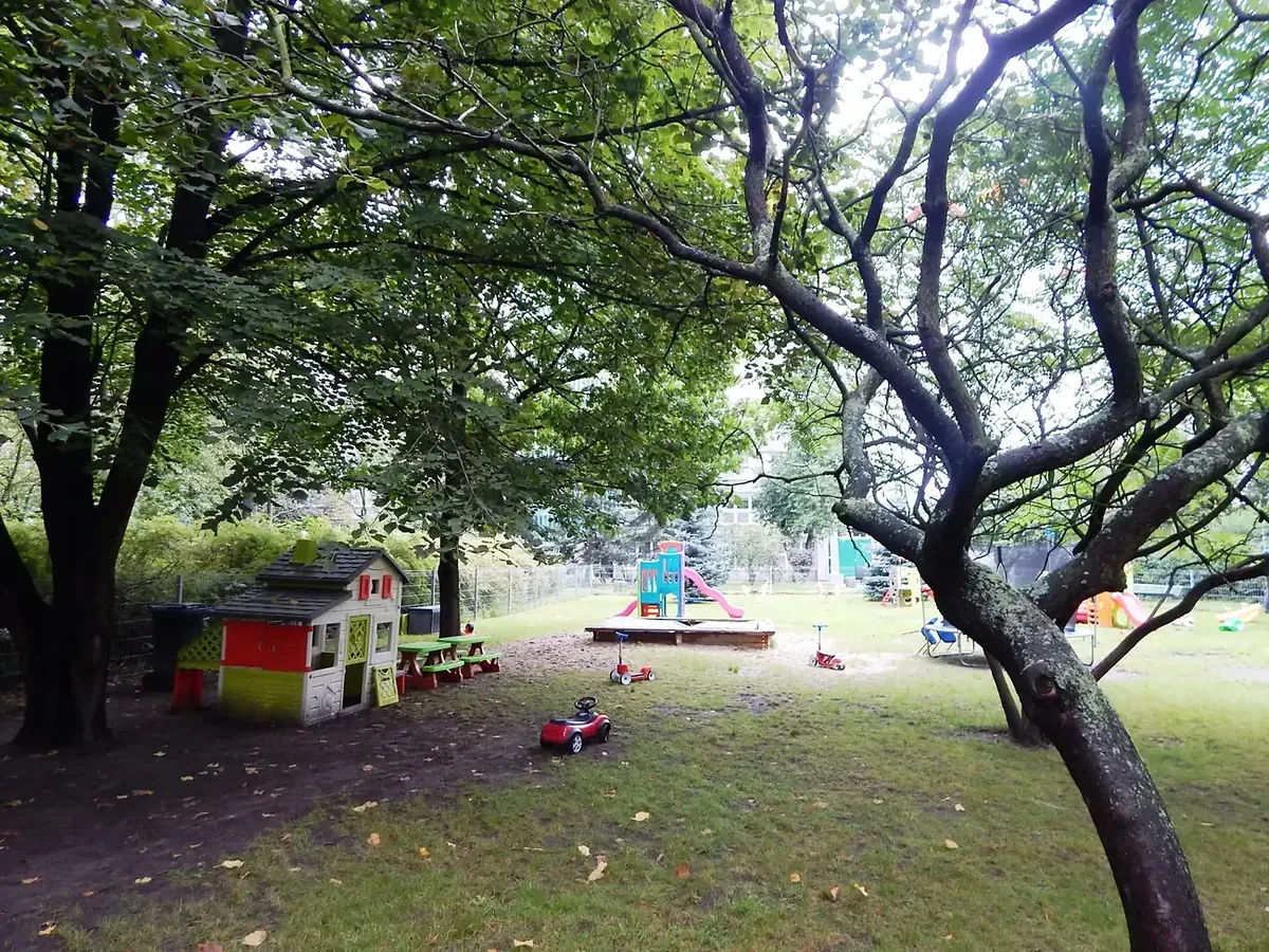 Duży, ogrodzony plac zabaw przedszkola wśród drzew